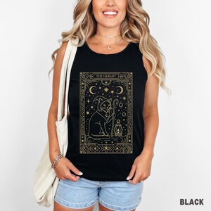 May include: Black tank top featuring a gold illustration of a cat in a wizard hat holding a staff and lantern. The text "THE HERMIT" is at the top of the design. The tank top is worn by a person wearing blue jean shorts.
