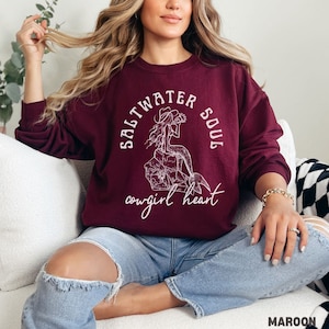woman wearing a maroon sweatshirt with a white design of a mermaid wearing a cowgirl hat and the phrase saltwater soul cowgirl heart