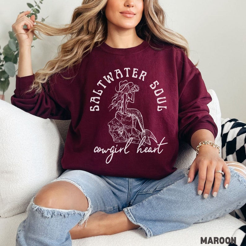 woman wearing a maroon sweatshirt with a white design of a mermaid wearing a cowgirl hat and the phrase saltwater soul cowgirl heart