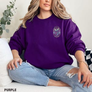 May include: A purple sweatshirt with a detailed mermaid tail design on the chest. The design features shades of blue, purple, and pink within an oval frame. The word "PURPLE" is printed at the bottom of the image.