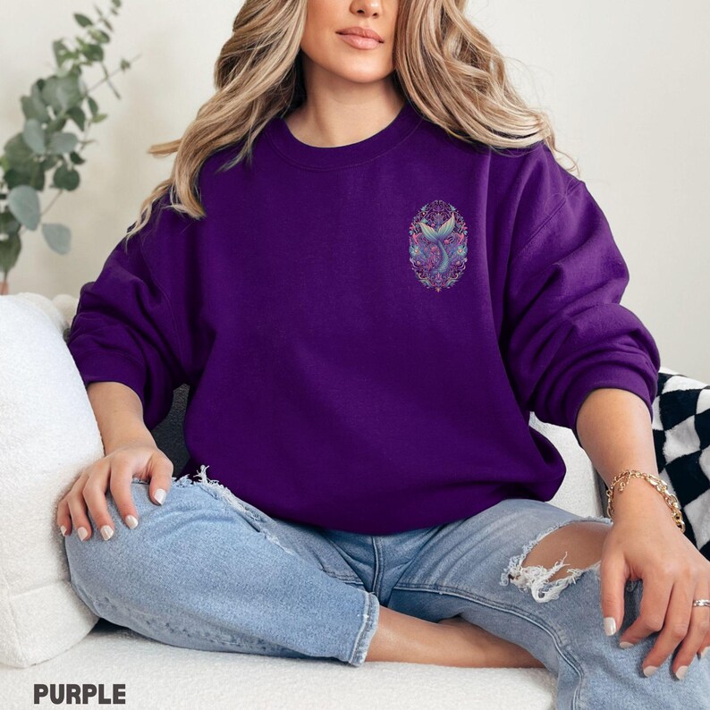 May include: A purple sweatshirt with a detailed mermaid tail design on the chest. The design features shades of blue, purple, and pink within an oval frame. The word "PURPLE" is printed at the bottom of the image.