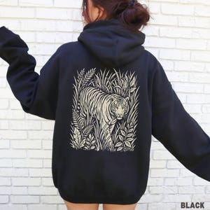 May include: Black hoodie with a cream-coloured tiger illustration and foliage on the back. The word "BLACK" is printed in white at the bottom right. The hoodie has a hood and long sleeves.