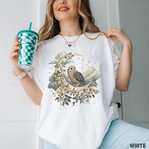 May include: White t-shirt featuring a detailed illustration of an owl perched on a crescent moon, surrounded by flowers, clouds, and stars. The design includes a celestial theme with a vintage aesthetic. The word "WHITE" is printed at the bottom.