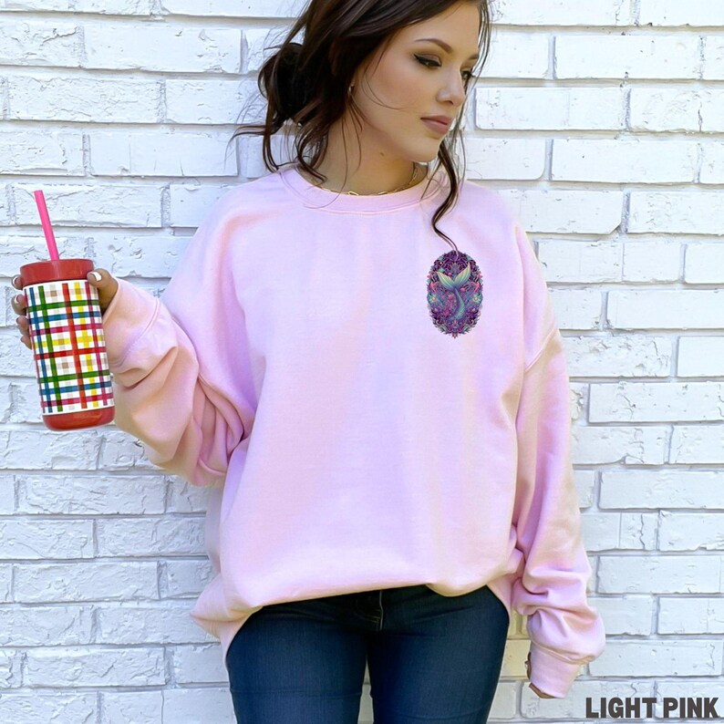 May include: Light pink sweatshirt with a mermaid graphic on the chest. The graphic features a mermaid tail and floral elements in shades of purple, blue, and green. The text "LIGHT PINK" is at the bottom right.