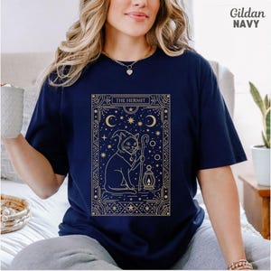 May include: Navy blue t-shirt with a gold graphic of a cat in a wizard hat holding a staff and lantern, with the text "THE HERMIT". The design is framed with stars and crescent moons.