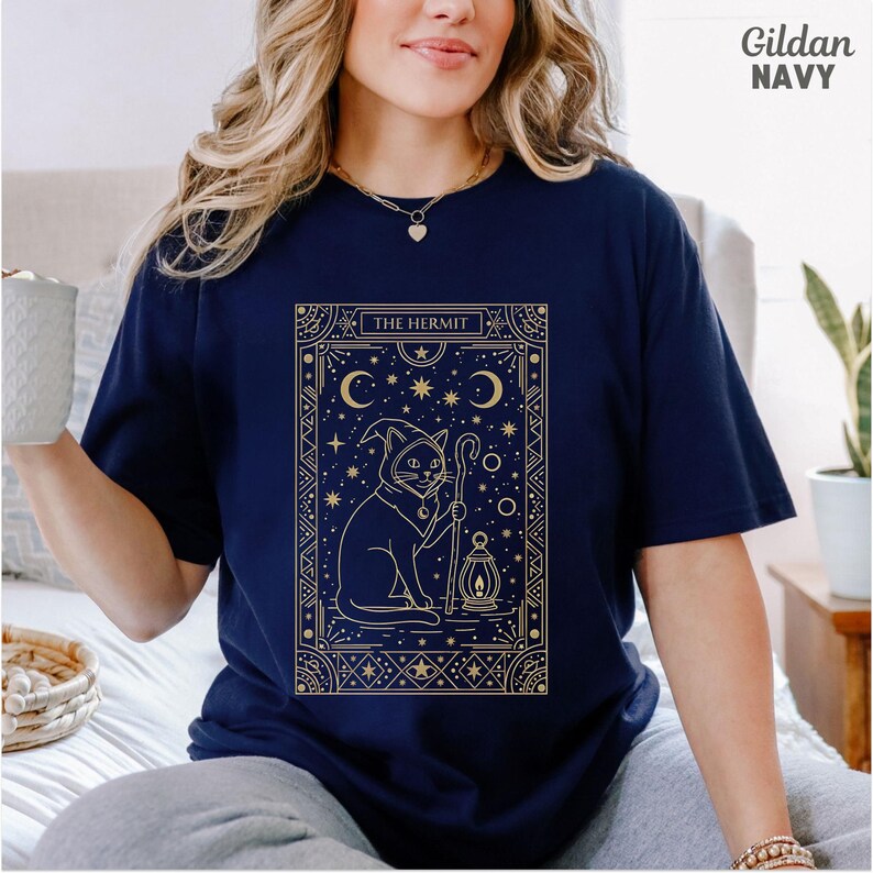 May include: Navy blue t-shirt with a gold graphic of a cat in a wizard hat holding a staff and lantern, with the text "THE HERMIT". The design is framed with stars and crescent moons.