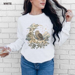 May include: White long-sleeve shirt with a celestial design. The graphic features an owl perched on a crescent moon, surrounded by flowers, clouds, and stars. The word "WHITE" is printed in the upper left corner.