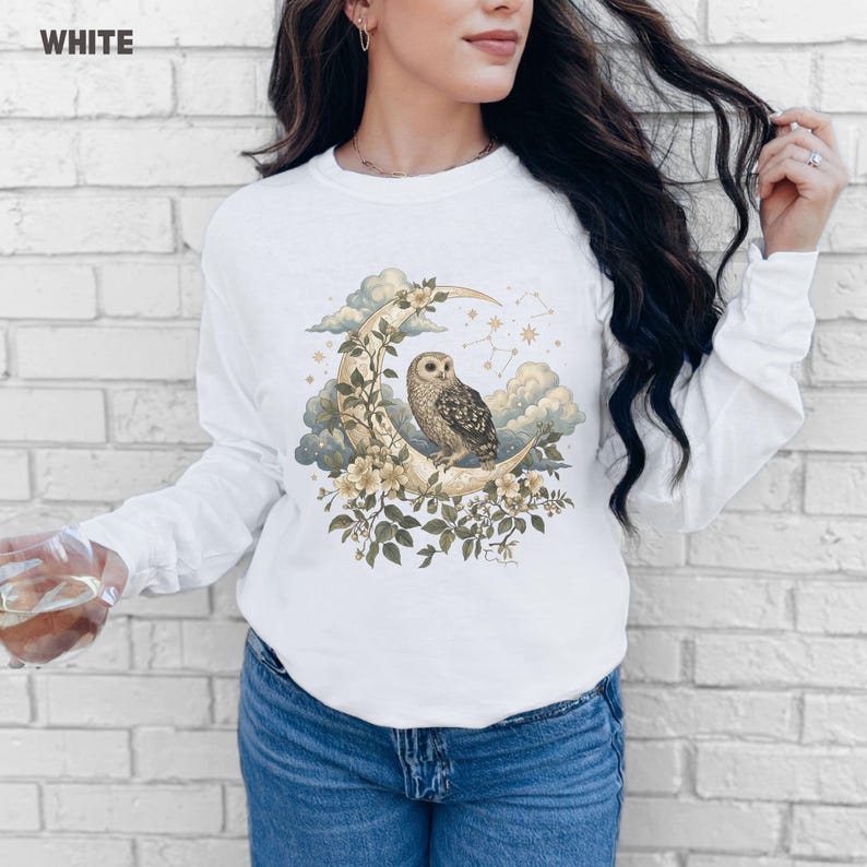 May include: White long-sleeve shirt with a celestial design. The graphic features an owl perched on a crescent moon, surrounded by flowers, clouds, and stars. The word "WHITE" is printed in the upper left corner.