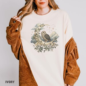 May include: Ivory-colored long-sleeved shirt featuring a detailed illustration of an owl perched on a crescent moon, surrounded by flowers, clouds, and stars. The shirt has a relaxed fit and is made of a soft material. The word "IVORY" is printed at the bottom.