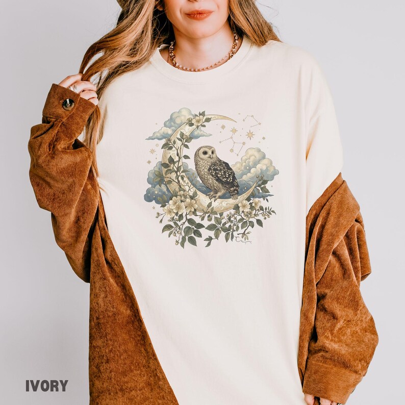 May include: Ivory-colored long-sleeved shirt featuring a detailed illustration of an owl perched on a crescent moon, surrounded by flowers, clouds, and stars. The shirt has a relaxed fit and is made of a soft material. The word "IVORY" is printed at the bottom.