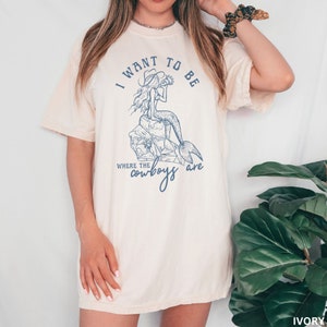 woman wearing a irovy shirt with a blue design showing a mermaid cowgirl and the phrase "i want to be where the cowboys are"