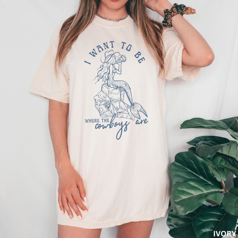 woman wearing a irovy shirt with a blue design showing a mermaid cowgirl and the phrase "i want to be where the cowboys are"