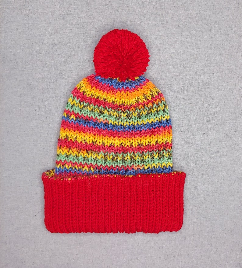 Double- layered multicolor beanie hat with red brim and bobble lying flat.