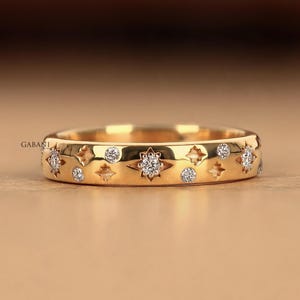 Könnte beinhalten: Ein goldener Bandring mit einer Reihe eingravierter Sternenmotive und kreisförmiger Fassungen. Einige der Sternmotive und kreisförmigen Vertiefungen sind mit kleinen, klaren, funkelnden Steinen besetzt. Das Wort GABANI ist links aufgedruckt.