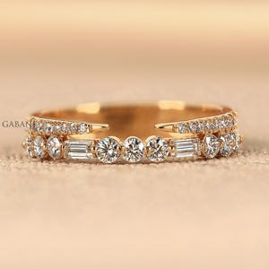 Puede incluir: Un anillo dorado con dos bandas. La banda superior está engastada con pequeñas piedras redondas y transparentes. La banda inferior presenta una fila de piedras más grandes, transparentes, redondas y de talla baguette. El anillo está sobre un fondo neutro.