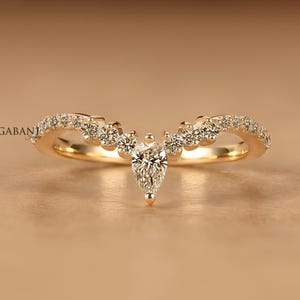 Könnte beinhalten: Ein goldener V-förmiger Ring mit einem zentralen Diamanten im Tropfenschliff und einer Reihe kleinerer Diamanten im Rundschliff entlang der Ringschiene. Das Schmuckstück zeigt links den Markennamen Gabani Jewelry und rechts unten ein Logo.