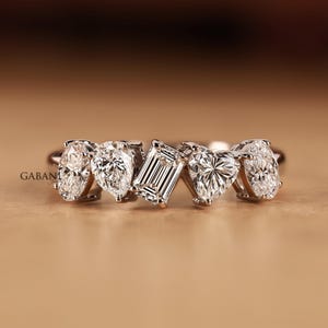 May include: A silver-toned ring featuring five distinct clear gemstones in a row. The stones are arranged in an oval cut, a pear cut, a central emerald cut, a heart cut, and a final oval cut, all set on a polished metallic band.