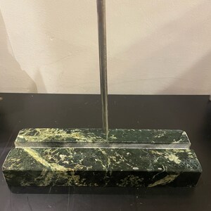 Può includere: Un porta biglietti da visita in marmo verde e bianco con un'asta verticale in metallo argentato. La base rettangolare ha un'incavatura per contenere le carte. Il marmo ha un motivo venato naturale.