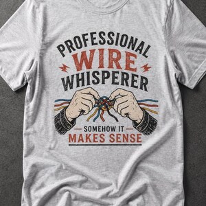 May include: A light gray heathered t-shirt featuring a graphic print of two hands untangling a cluster of colorful wires. The text reads PROFESSIONAL WIRE WHISPERER and SOMEHOW IT MAKES SENSE, accented with small red lightning bolt icons.