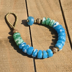 May include: A handmade bracelet featuring blue, green, and white beads. The bracelet has a loop closure and decorative gold-colored metal accents. The beads are a mix of round and flat shapes, creating a unique design.
