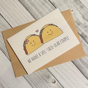May include: A white card with a brown envelope. The card has a drawing of two tacos with faces. The text on the card reads "We make a spec-taco-ular couple".