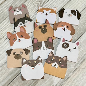 May include: A collection of die-cut paper dog faces in various breeds, including a pug, husky, and corgi. Each dog has a unique expression and color scheme, with shades of brown, white, and black. The dog faces are arranged on a light wood-grain surface.
