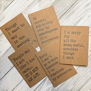May include: Five brown kraft paper notebooks with inspirational quotes printed in black text. The quotes are: "Things I want to say in the meeting", "People say I act like I don't care... It's not an act.", "Be careful not to trip over my amazingness; it's everywhere.", "Sorry I can't go to work tomorrow I've fractured my motivation.", and "I'm sorry for all the mean, awful, accurate things I said."
