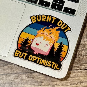 May include: A round sticker with a cartoon marshmallow on fire, with the text "BURNT OUT BUT OPTIMISTIC". The design features a sunset and pine trees. The sticker is on a laptop.