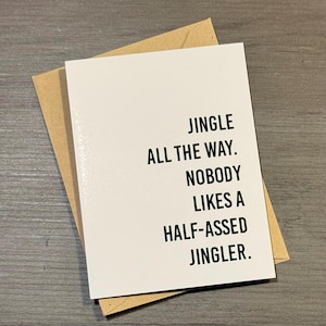 May include: A white greeting card with black text that reads "JINGLE ALL THE WAY. NOBODY LIKES A HALF-ASSED JINGLER." The card is inside a brown envelope, set against a gray wooden surface.