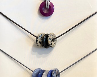 May include: Three necklaces with different colored stone beads. The top necklace has a single purple stone bead. The middle necklace has three beads: one black, one white with black spots, and one white with black spots. The bottom necklace has four beads: two blue, one white, and one blue.