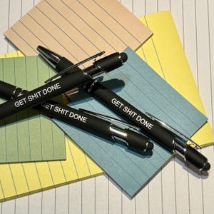 May include: Black pens with silver accents and the phrase "GET SHIT DONE" printed in white. The pens are lying on top of lined paper in shades of blue, yellow, and tan.