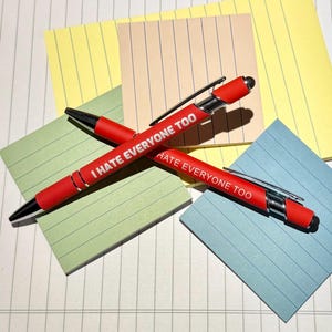 May include: Two red pens with the text "I HATE EVERYONE TOO" printed in white. The pens are lying on a white surface with colorful sticky notes in the background.