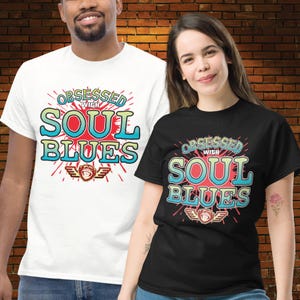 Könnte beinhalten: Zwei T-Shirts, eines weiß und eines schwarz, mit dem Text "Obsessed with Soul Blues" in einem farbenfrohen Design. Das Design beinhaltet einen Affen mit Flügeln. Die Shirts sind kurzärmlig.