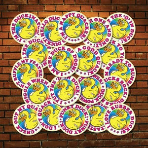 May include: A selection of circular stickers with a yellow cartoon duck and a rainbow border. Each sticker has a different phrase, including "I'm the Duckinator!" and "Happy Quacker!" The stickers are displayed against a brick wall.