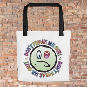 Könnte beinhalten: Weiße Tragetasche mit schwarzen Griffen. Auf der Tasche ist ein Cartoon-Gesicht mit dem Text "DON'T FREAK ME OUT!" in Regenbogenfarben abgebildet. Das Gesicht hat ein blaues Auge, ein Spiralauge und einen traurigen Mund.
