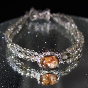 Könnte beinhalten: Ein silbernes Armband mit einem klaren Kristall und einem braunen Edelstein, der in einer silbernen Fassung sitzt. Das Armband besteht aus mehreren Reihen von Perlen.