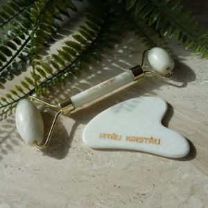 May include: White jade facial roller and gua sha tool with gold accents. Both tools have the text "VITAU KRISTALU" engraved on them.