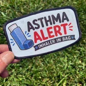 May include: A rectangular embroidered patch with a white background and a black border. It features a blue inhaler illustration, the black text ASTHMA, the red text ALERT, and a black banner with the white text INHALER IN BAG and blue hearts.