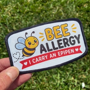 May include: A rectangular patch with a white background and a black border. It features a cartoon bee, the yellow text BEE ALLERGY, and a red banner with the white text I CARRY AN EPIPEN, flanked by two small white hearts on the banner.