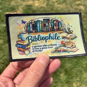 May include: A rectangular fabric patch with a cream background featuring a colorful illustration of books, a coffee mug, a sleeping cat, and a candle. The text reads: Bibliophile noun: a person who collects or has a great love of books.