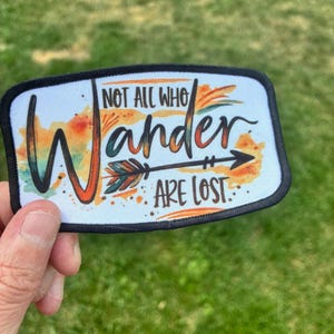 May include: A rectangular fabric patch with a black stitched border featuring a white background with watercolor-style orange, yellow, and teal splashes. The text reads "NOT ALL WHO wander ARE LOST" with an arrow graphic integrated into the design.