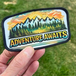 May include: A rectangular embroidered patch with a black border featuring a colorful landscape of snow-capped mountains, a dense green pine forest, and a bright orange and yellow sunset sky. The text ADVENTURE AWAITS is printed in bold.