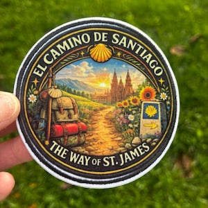 May include: A circular embroidered patch featuring a landscape illustration of a hiking trail leading to a cathedral at sunrise. The text reads EL CAMINO DE SANTIAGO and THE WAY OF ST. JAMES. It includes a backpack, a scallop shell, and sunflowers.
