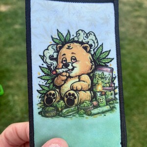 May include: A rectangular fabric patch with a black stitched border featuring a cartoon illustration of a light brown teddy bear smoking a joint, surrounded by green cannabis leaves, a jar labeled Nubs, a green lighter, and loose plant material.