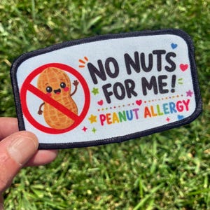 May include: A rectangular patch with a white background and a black border. It features a cartoon peanut inside a red prohibition circle, with the text NO NUTS FOR ME! and PEANUT ALLERGY in colorful letters, surrounded by small hearts and stars.