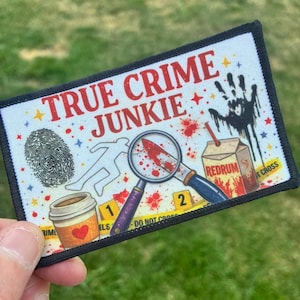 May include: A rectangular embroidered patch with a white background and black border. It features the text TRUE CRIME JUNKIE in red, a black fingerprint, a white chalk outline, a magnifying glass, a bloody knife, and a carton labeled REDRUM.