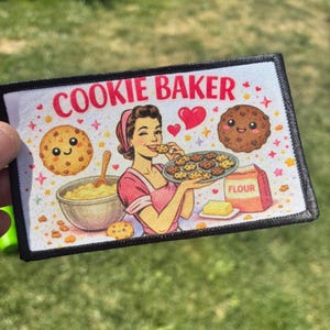May include: A rectangular fabric patch with a black border featuring the text COOKIE BAKER in red. The illustration shows a person in a pink apron holding a tray of cookies, a mixing bowl, a bag of flour, and two smiling cookie characters.