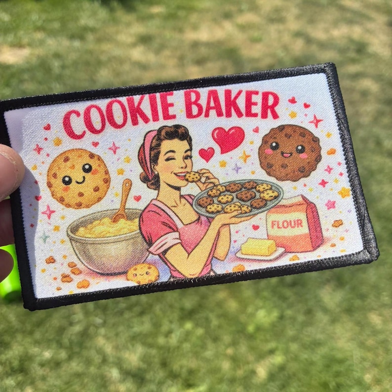 A rectangular fabric patch with a black border featuring the text COOKIE BAKER in red. The illustration shows a person in a pink apron holding a tray of cookies, a mixing bowl, a bag of flour, and two smiling cookie characters.