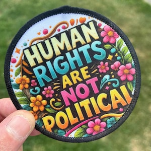 May include: A circular fabric patch with a black stitched border featuring the text "HUMAN RIGHTS ARE NOT POLITICAL" in bold, colorful letters. The design is decorated with vibrant orange, yellow, and pink flowers and green leaves.
