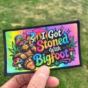 May include: A rectangular fabric patch with a tie-dye background featuring a brown Bigfoot wearing a colorful beanie and sunglasses, holding a smoking item. The text reads I Got Stoned With Bigfoot, surrounded by cannabis leaves and mushrooms.